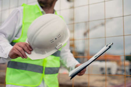 Chief in a vest and a construction helmet with documents in his hands, against the background of a tower crane and the sky with clouds. House construction inspectionの写真素材