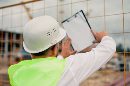 Chief in a vest and a construction helmet with documents in his hands, against the background of a tower crane and the sky with clouds. House construction inspectionの写真素材