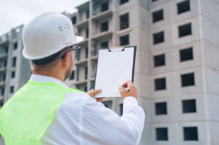 Chief in a vest and a construction helmet with documents in his hands, against the background of a tower crane and the sky with clouds. House construction inspectionの写真素材