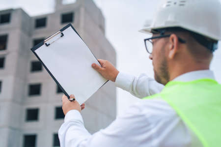 Chief in a vest and a construction helmet with documents in his hands, against the background of a tower crane and the sky with clouds. House construction inspectionの写真素材