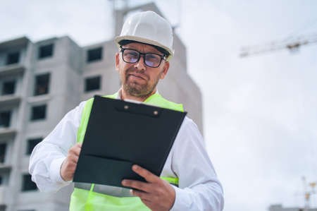 Chief in a vest and a construction helmet with documents in his hands, against the background of a tower crane and the sky with clouds. House construction inspectionの写真素材