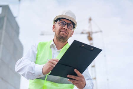 Chief in a vest and a construction helmet with documents in his hands, against the background of a tower crane and the sky with clouds. House construction inspectionの写真素材