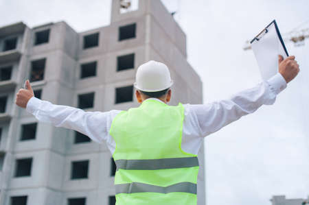 Chief in a vest and a construction helmet with documents in his hands, against the background of a tower crane and the sky with clouds. House construction inspectionの写真素材