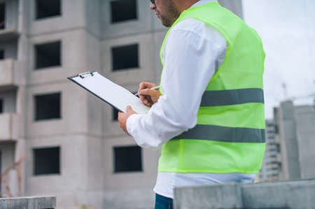 Chief in a vest and a construction helmet with documents in his hands, against the background of a tower crane and the sky with clouds. House construction inspectionの写真素材
