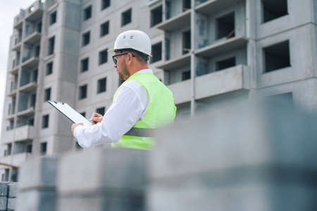 Chief in a vest and a construction helmet with documents in his hands, against the background of a tower crane and the sky with clouds. House construction inspectionの写真素材