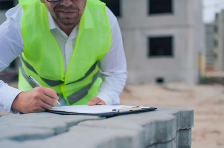 Chief in a vest and a construction helmet with documents in his hands, against the background of a tower crane and the sky with clouds. House construction inspectionの写真素材
