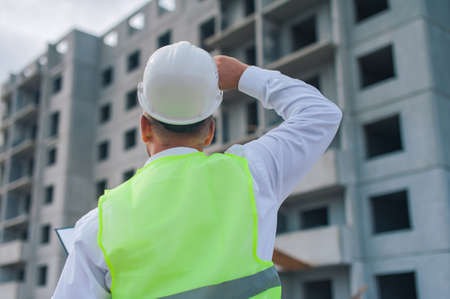 Chief in a vest and a construction helmet with documents in his hands, against the background of a tower crane and the sky with clouds. House construction inspectionの写真素材