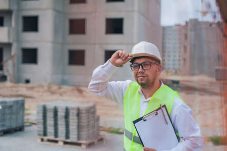 Chief in a vest and a construction helmet with documents in his hands, against the background of a tower crane and the sky with clouds. House construction inspectionの写真素材