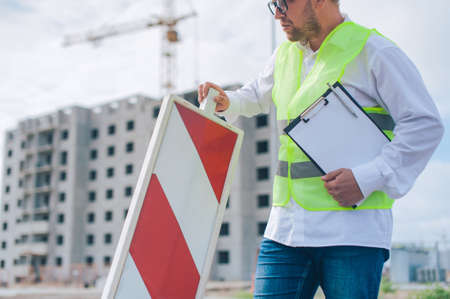 Chief in a vest and a construction helmet with documents in his hands, against the background of a tower crane and the sky with clouds. House construction inspectionの写真素材