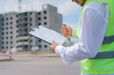 Chief in a vest and a construction helmet with documents in his hands, against the background of a tower crane and the sky with clouds. House construction inspectionの写真素材