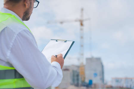 Chief in a vest and a construction helmet with documents in his hands, against the background of a tower crane and the sky with clouds. House construction inspectionの写真素材