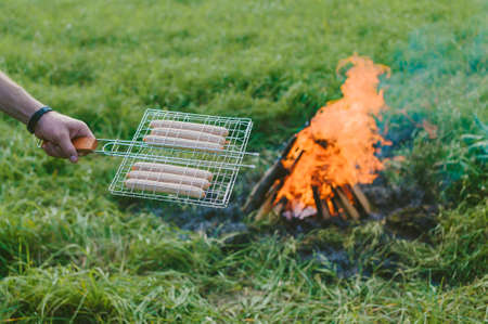 A man with a grate in his hand prepares sausages on the fire. Picnic in natureの写真素材