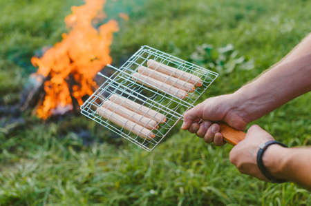 A man with a grate in his hand prepares sausages on the fire. Picnic in natureの写真素材