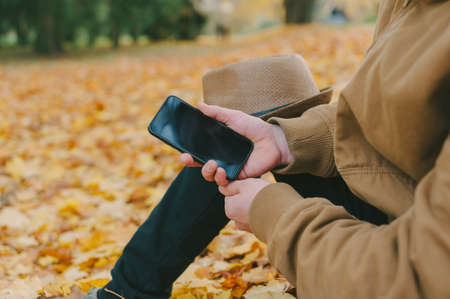 A man in a hat with a smartphone on an autumn day against a background of beautiful leavesの写真素材