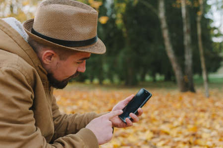 A man in a hat with a smartphone on an autumn day against a background of beautiful leavesの写真素材