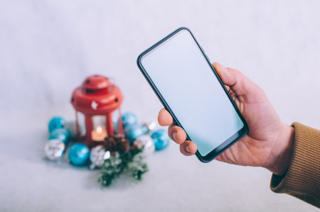 The guy holds a smartphone with a white screen in his hands on the background of a Christmas compositionの写真素材
