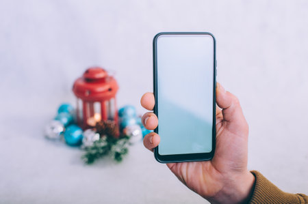 The guy holds a smartphone with a white screen in his hands on the background of a Christmas compositionの写真素材