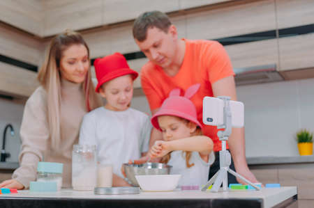 Mom with Father teaches two daughters to cook dough in the kitchen. The family is filming a culinary video on a smartphoneの写真素材