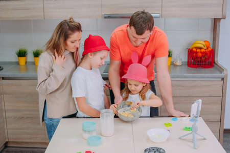 Mom with Father teaches two daughters to cook dough in the kitchen. The family is filming a culinary video on a smartphoneの写真素材