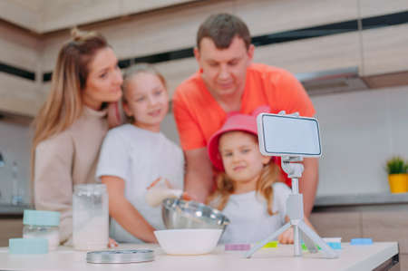 Mom with Father teaches two daughters to cook dough in the kitchen. The family is filming a culinary video on a smartphoneの写真素材
