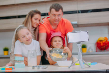 Mom with Father teaches two daughters to cook dough in the kitchen. The family is filming a culinary video on a smartphoneの写真素材