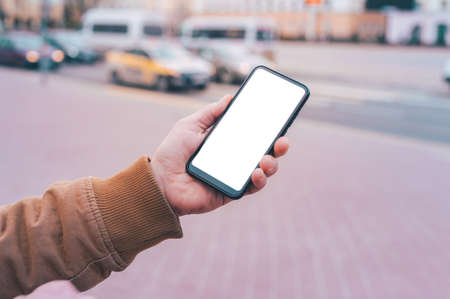 A man holds a mock-up of a smartphone with a white screen on the background of the roadの写真素材