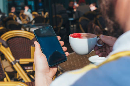Man holds a smartphone on the background of a cup of coffee in a restaurantの写真素材