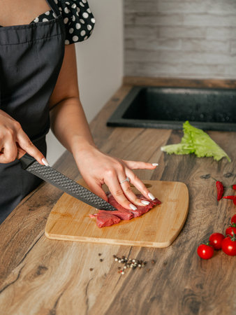 Woman cuts meat with a knife on a wooden board. Kitchen location.