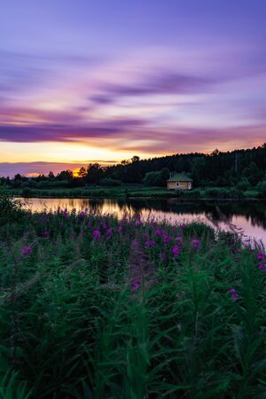 Beautiful long exposure landscape at sunset with purple sky and flowers and lonely little house at the bank of small pondの写真素材
