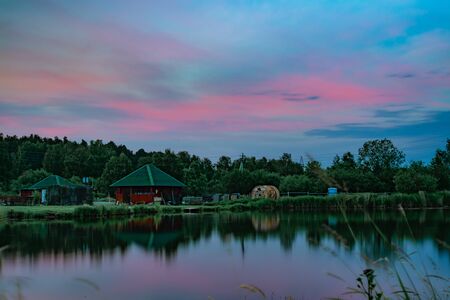 Long exposure scenic view of small red house on the bank of still pond reflecting beautiful blue and...