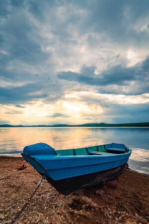 Blue boat moored on the shore of lake under cloudy sky at sunsetの写真素材