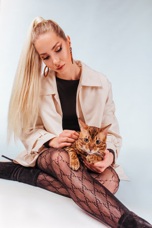 Portrait of an attractive cute young blonde woman with a bengal cat. A beautiful girl sitting on the floor looks at the cat, elegant hairstyle, cheeky makeup, blue eyes. The background is white.の写真素材