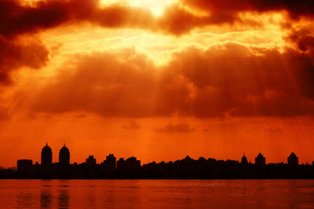 City silhouette and red sky with sun rays through the clouds  River water with reflection on foregroundの写真素材