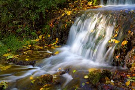small waterfall with autumn leaves in flowing waterの写真素材