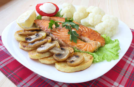 Fried salmon with potatoes, mushrooms, cauliflower, tomato and lemon on white plateの写真素材