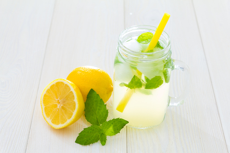 Fancy cool glass of lemonade with ice and mint, Mason jar style cup with yellow straw, green leaves of fresh mint and lemons, one of lemon cut in half. Refreshment still life on white tableの写真素材