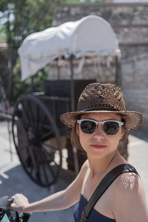 The young woman in the sunglasses and the straw hat is photographed against the background of the wooden cart during the walkの写真素材