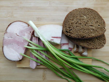 Close up view slices of smoked bacon meat with rye whole grain black bread, green onion and pepper on the white table. Selective focus, copy space. Country style organic food.の写真素材