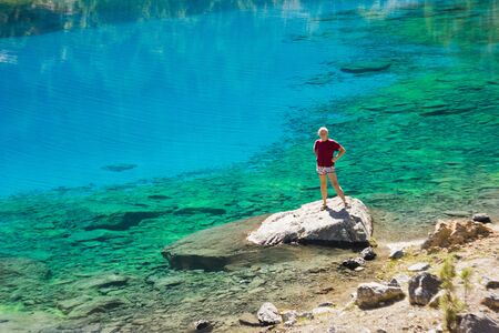 girl in red t-short near deep blue lake standing and smiling on a stoneの写真素材