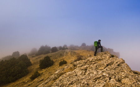 man with green backpack walking on the edge of a cliff in foggy weatherの写真素材