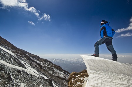 happy man in blue jacket with hands-up standing on a cliff in mountains at sunriseの写真素材