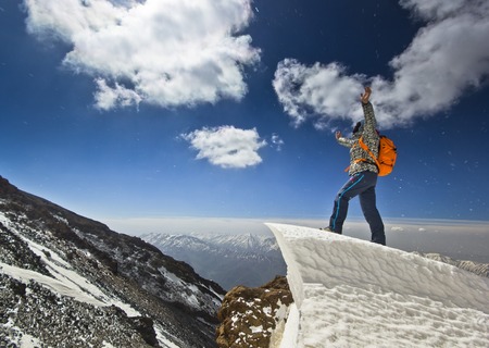 happy man with orange backpack with hands-up standing on a cliff in mountains at sunriseの写真素材