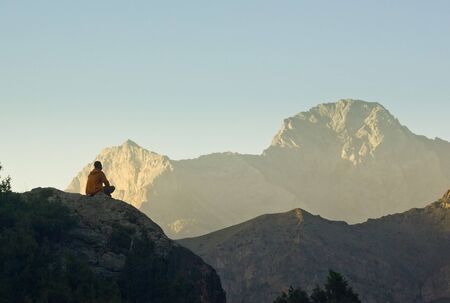 man sitting and meditating on a cliff at sunset surrounded by high rocky mountainsの写真素材