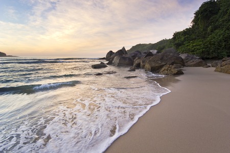 purple sunrise in tropical beach with water waves, sand and stones on shoreの写真素材