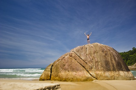 man with hands up standing on big stone in tropical beach with green mountains and huge waves on waterの写真素材