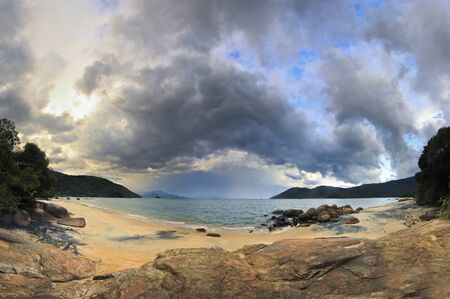 panorama of sunset with big clouds in tropical beach with water waves, sand and stones on shoreの写真素材