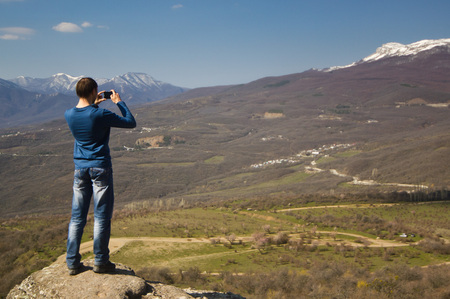 man in casual clothes on a  mountain cliff and making photo by phoneの写真素材