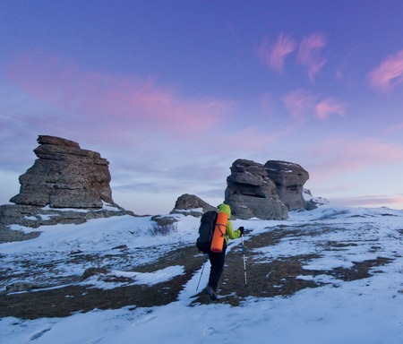 man with backpack and poles going in mountains and rocks snow covered at purple sunsetの写真素材