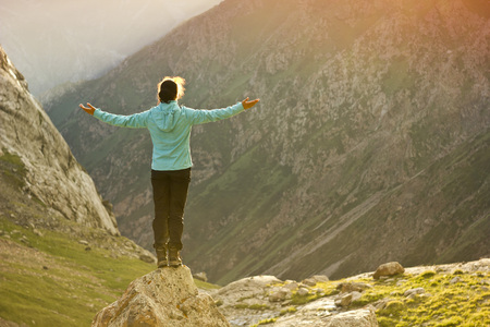 girl in blue jacket standing on stone with hands-up above mountais at sunsetの写真素材