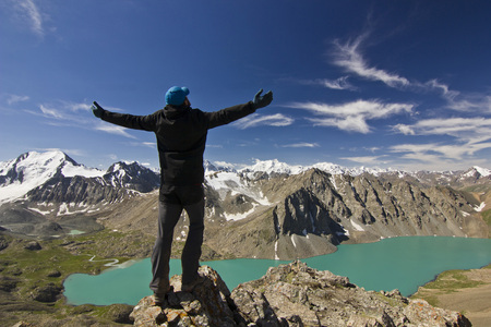 man in black jacket standing with hands up above blue calm mountain lake Alakol with mountains surroundedの写真素材